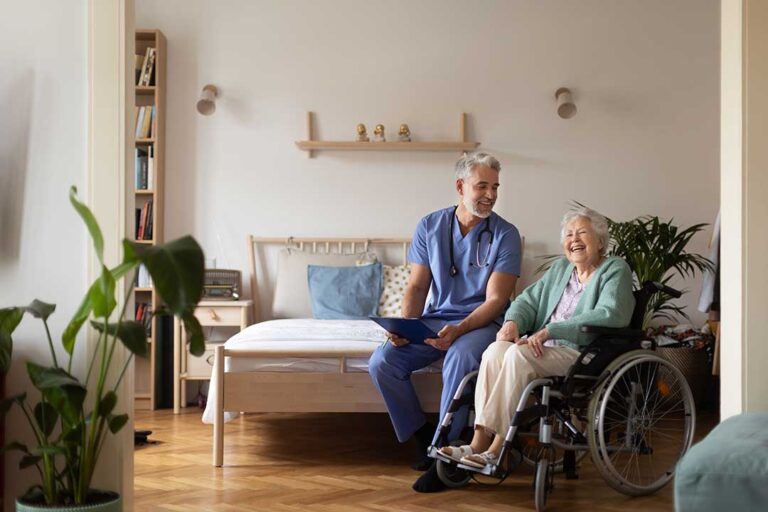 Older adults share a meal while a caregiver assists a resident in a bright community dining room.